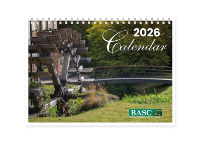 Cover of 2026 calendar featuring an outdoor scene with a wooden waterwheel, a modern pedestrian bridge, lush greenery, and partially visible buildings. '2026 Calendar' text in white script at the top right, with the BASC logo at the bottom right