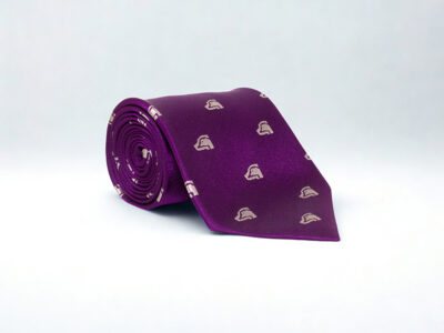 A deep purple silk tie, featuring the BASC masthead logo embroidered in silver, neatly rolled up on a white background. 