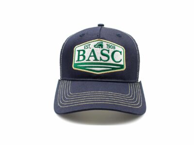 Front view of a navy blue baseball cap with contrast stitching on the peak and an embroidered BASC badge in green, beige, and white reading “EST. 1908”.